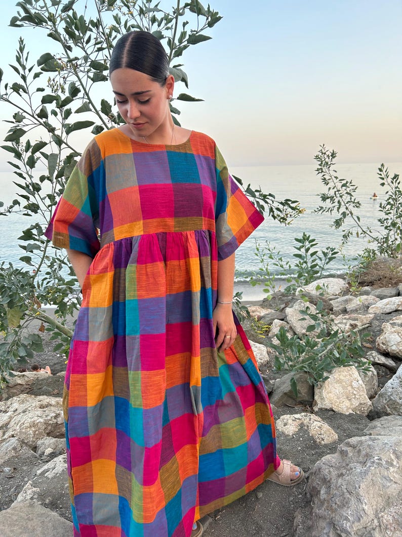 May include: A colorful, patterned dress with a square neckline and short sleeves. The dress features a vibrant plaid design in shades of orange, pink, purple, blue, and green. The dress is worn outdoors.