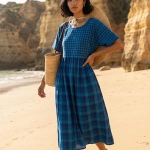 May include: A blue plaid dress with short sleeves and a gathered waist. The dress is knee-length and worn with a straw tote bag. The background shows a sandy beach and cliffs.