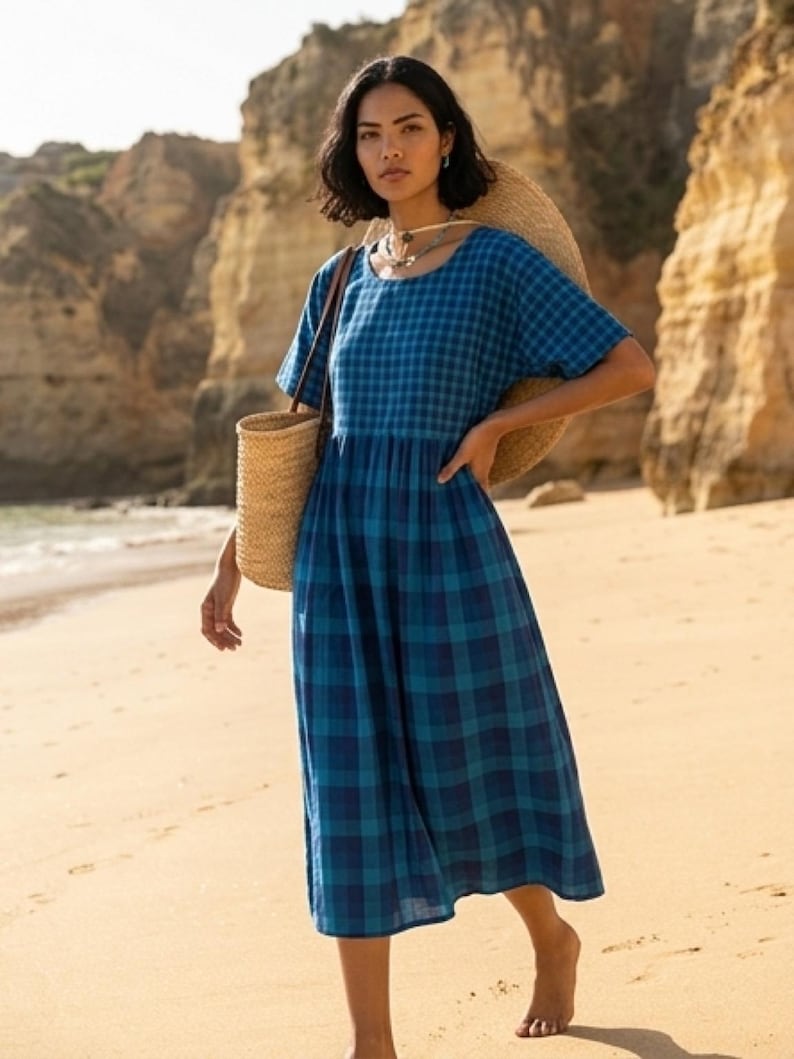 May include: A blue plaid dress with short sleeves and a gathered waist. The dress is knee-length and worn with a straw tote bag. The background shows a sandy beach and cliffs.
