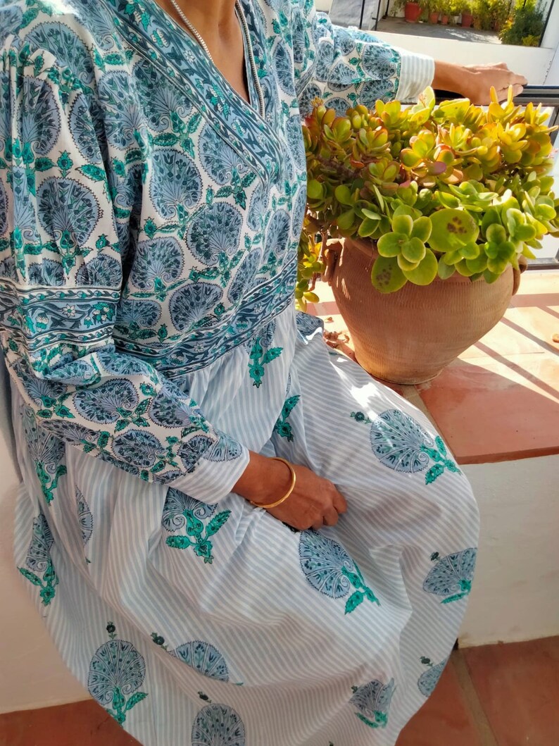 May include: A long, white dress with blue and green floral print. The dress has a V-neck and long sleeves. The dress is made of a lightweight fabric and has a flowing silhouette.