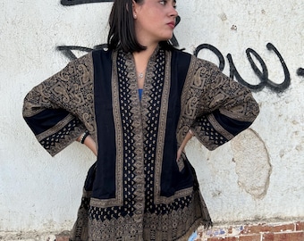 Puede incluir: Chaqueta negra y dorada con estampado de cachemira y ribete de flecos. La chaqueta presenta un diseño de paneles verticales con un patrón detallado y un borde contrastante. Las mangas y el dobladillo muestran el intrincado diseño.