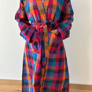 Könnte beinhalten: Ein langer, farbenfroher Bademantel mit einem Karomuster in Rot-, Orange-, Lila-, Blau- und Grüntönen. Der Bademantel hat einen V-Ausschnitt, lange Ärmel, Taschen und einen passenden Gürtel. Der Bademantel ist aus leichtem Stoff.