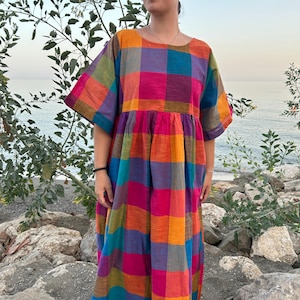May include: A colorful, long, plaid dress in shades of pink, orange, blue, green, and purple. The dress has short sleeves and a loose, flowing fit. The model is standing outdoors near the water.