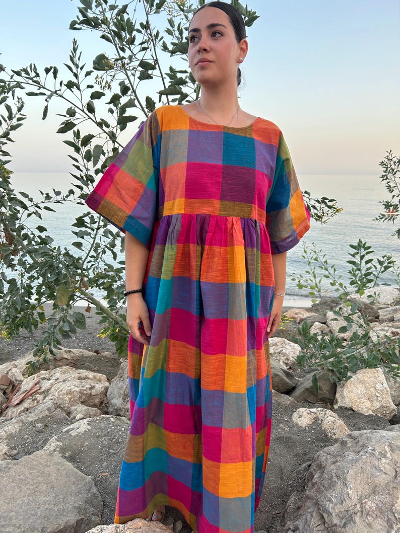 May include: A colorful, long, plaid dress in shades of pink, orange, blue, green, and purple. The dress has short sleeves and a loose, flowing fit. The model is standing outdoors near the water.