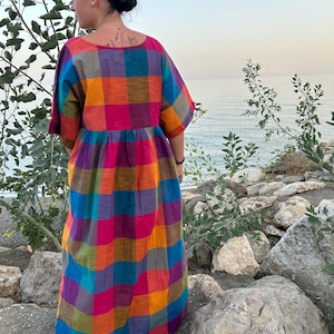 May include: A long, colorful plaid dress in shades of pink, orange, blue, and purple. The dress has short sleeves and a loose fit. The person is standing outdoors near the water.