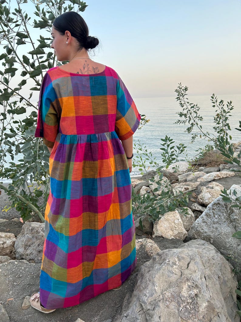 May include: A long, colorful plaid dress in shades of pink, orange, blue, and purple. The dress has short sleeves and a loose fit. The person is standing outdoors near the water.