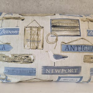 May include: A rectangular throw pillow featuring a nautical-themed print on a cream background. The design includes blue and tan wooden signs with text such as Devon, Perth, Antigua, and Newport, along with a seagull illustration and rope.