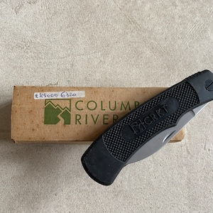 May include: A black folding knife with a black handle and a silver blade. The knife is closed and has the text "CRKT" on the handle. The knife is sitting on top of a brown cardboard box with the text "COLUMBIA RIVER" on it.