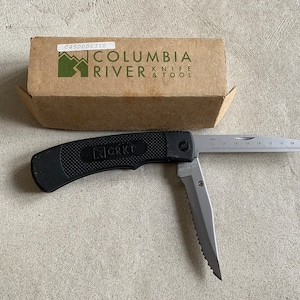 May include: A black folding knife with a serrated blade and a ruler on the back. The knife is open and the blade is extended. The knife is made by CRKT and is packaged in a brown box with the Columbia River Knife & Tool logo.
