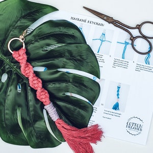 May include: A pink and white macrame keychain with a tassel. The keychain is made using a knotting technique and is hanging from a gold key ring. The image also includes instructions for making a macrame keychain.