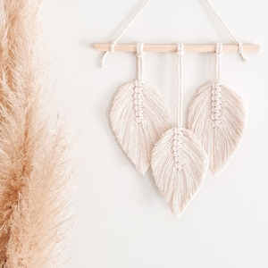 May include: A macramé wall hanging with three cream-coloured leaf designs suspended from a natural wood dowel. The leaves are intricately knotted, showcasing a textured, bohemian style. The wall decor is hung against a white wall, with dried pampas grass in the background.