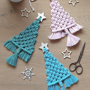 May include: Three macrame Christmas tree decorations in teal, light pink, and light blue, each with a wooden star topper and tassel base. Additional wooden stars, a snowflake, and scissors are scattered around.