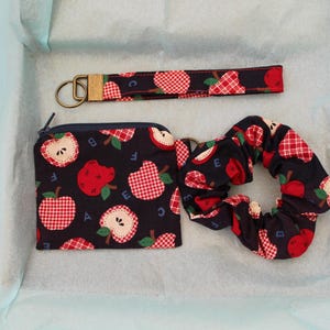 Peut inclure: Un ensemble en tissu avec un fond noir, un motif de pommes à carreaux rouges et blancs et des lettres bleues. L'ensemble comprend une pochette rectangulaire zippée, un chouchou circulaire et un porte-clés dragonne avec un anneau en métal doré.