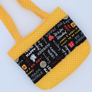 May include: A yellow tote bag with white polka dots featuring a central black fabric panel printed with white and red Mickey Mouse themed text and icons including phrases like 100% HAPPY, OH BOY, and M is for Mischief with small red mouse ears.