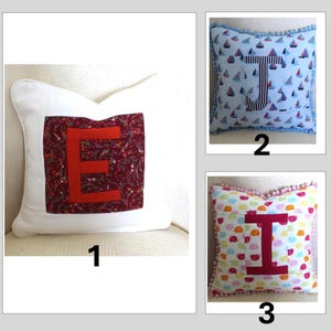 May include: Three square throw pillows featuring centered fabric letters. Pillow 1 is white with a maroon floral square and red letter E. Pillow 2 has a blue sailboat pattern, a striped letter T, and blue pom-pom trim. Pillow 3 has colorful elephants, a pink letter I, and pink pom-pom trim.