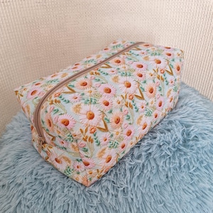 Peut inclure: Une trousse rectangulaire zippée avec une base en tissu vert menthe clair, ornée d'un motif brodé de marguerites roses et orange, accompagnées de tiges et de feuilles vertes. La trousse présente une fermeture éclair beige sur le dessus.