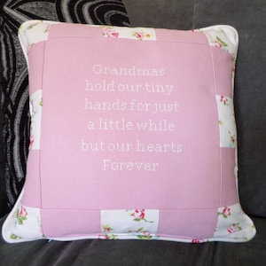 Op de afbeelding: Een roze en wit patchwork kussen met een wit geborduurd citaat: "Oma's houden onze kleine handjes vast voor een korte tijd, maar onze harten voor altijd".