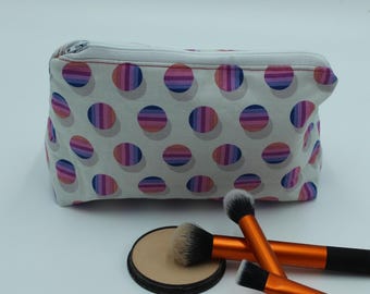 May include: A rectangular zippered pouch featuring a white background with a repeating pattern of multicolored striped circles in shades of pink, purple, and blue. Two makeup brushes with orange handles and a round compact are positioned in front.