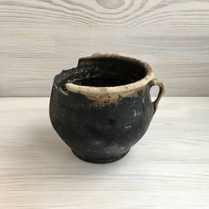 Puede incluir: Una olla de cerámica rústica, hecha a mano, con un exterior oscuro y texturizado y un borde de color claro. La olla tiene un asa pequeña y una forma irregular única, lo que sugiere que es una pieza antigua o vintage.