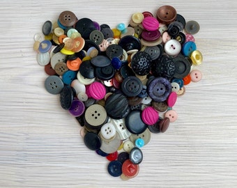 May include: A heart shape composed of various buttons in different colors and sizes. The buttons include shades of black, brown, pink, white, and blue. Some buttons have intricate designs, while others are plain.