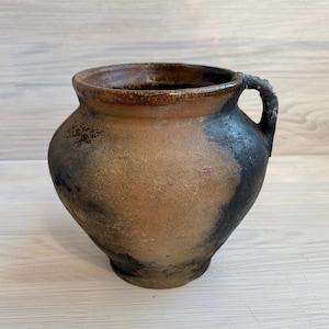May include: A rustic earthenware pot with a wide, rounded body and a single side handle. The surface features a textured, mottled finish with blended tones of earthy brown, tan, and deep charcoal black, showing a weathered, uneven appearance.