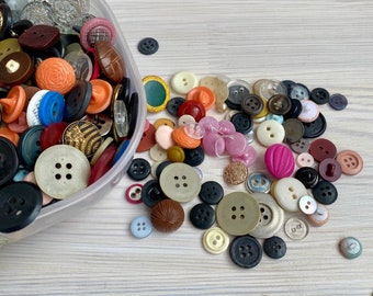 May include: A clear plastic container overflowing with a variety of buttons in various sizes, shapes, and colors. Buttons are scattered on a light wood-grain surface. Colors include black, white, orange, pink, and brown.