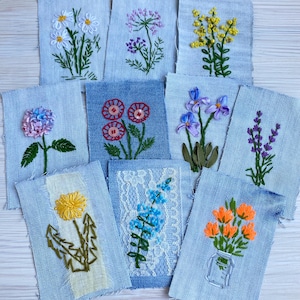 May include: A collection of nine rectangular denim fabric patches featuring various floral embroidery designs, including daisies, lavender, poppies with button centers, and other botanical motifs, some accented with white lace or yellow fabric.