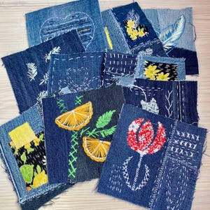 May include: A collection of denim fabric patches, each uniquely embroidered with various designs. The patches feature images of flowers, leaves, and citrus fruits, stitched with white, yellow, and green thread. The denim is a mix of light and dark blue.