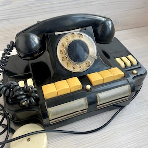Puede incluir: Un teléfono negro vintage con un dial rotatorio y un auricular. El teléfono tiene una serie de botones amarillos y un altavoz. El diseño del teléfono es retro, con una estética clásica. El teléfono está conectado a una toma de corriente con un cable negro.