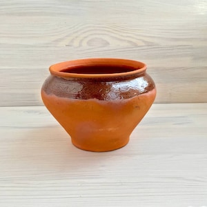 Peut inclure: Un pot en terre cuite brun avec une large ouverture et un corps arrondi. Le pot est recouvert d'un glacis brun avec un motif décoratif autour du haut.