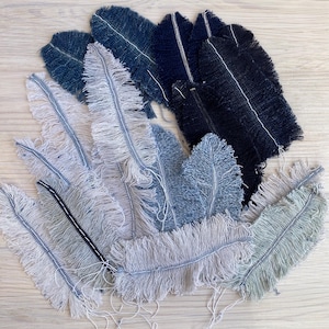 May include: A collection of handmade fabric feathers in various shades of blue, gray, and black. Each feather has a frayed edge and a central stitched line. The feathers are arranged on a light wooden surface.