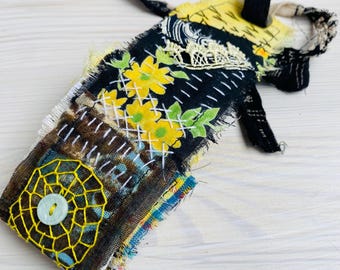May include: A handmade fabric spray bottle cover with a patchwork design. The cover features yellow floral embroidery on a black background, a yellow web design with a button, and white stitching. The cover is made of various fabrics.