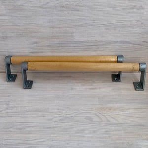 Può includere: Due maniglie per mobili in legno con finitura naturale e staffe metalliche. Le maniglie sono cilindriche e marrone chiaro, con staffe metalliche grigie ad ogni estremità per il montaggio. Sfondo con venature del legno chiaro.