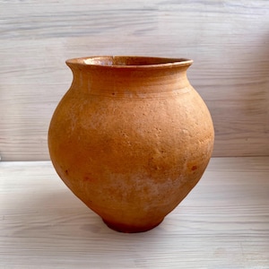 Puede incluir: Un jarrón de cerámica de color terracota, con un cuerpo redondeado y un borde ligeramente ensanchado. El recipiente hecho a mano tiene una superficie texturizada y un tono cálido y terroso, lo que sugiere una estética vintage o antigua. El jarrón se muestra sobre un fondo de madera clara.
