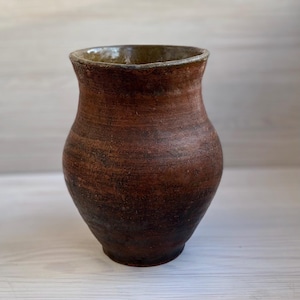 Könnte beinhalten: Eine rustikale, handgefertigte braune Keramikvase mit breitem Körper und leicht ausgestelltem Rand. Die Vase hat eine strukturierte Oberfläche und eine dunkle, erdige Farbe, perfekt für die Präsentation von Blumen oder als dekoratives Stück. Die Vase ist etwa 20 cm hoch.