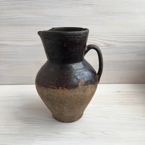 Puede incluir: Una jarra de cerámica marrón y beige con asa. La jarra tiene un aspecto rústico y vintage.