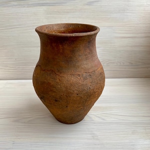 Peut inclure: Un vase en terre cuite brun rustique, avec un corps large et arrondi et un bord évasé. La surface présente un aspect texturé et vieilli, avec des fissures visibles et des variations de couleur. Le vase est fait à la main.