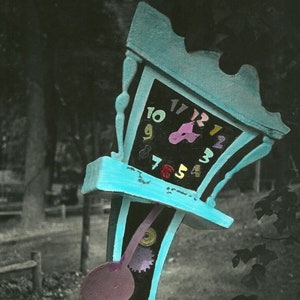 May include: A whimsical, blue, wooden clock with a colorful face and a large, purple pendulum. The clock is tilted on its side and appears to be broken. The clock is sitting on a wooden bench with a blue paint finish.