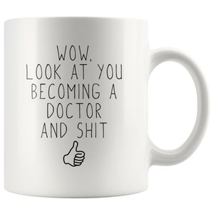 May include: White ceramic coffee mug with black text that reads "WOW, LOOK AT YOU BECOMING A DOCTOR AND SHIT" with a thumbs up icon.