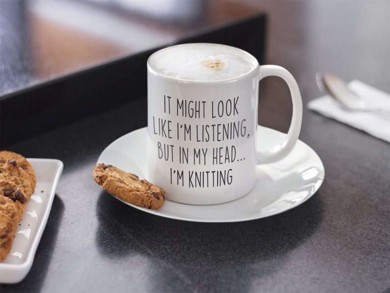 May include: White ceramic mug with a black text that reads "It might look like I'm listening, but in my head... I'm knitting". The mug is filled with a light brown liquid and has a white saucer. There are chocolate chip cookies on a white plate next to the mug.