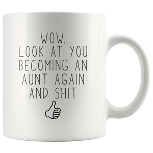 May include: White ceramic coffee mug with black text that reads "WOW, LOOK AT YOU BECOMING AN AUNT AGAIN AND SHIT" with a thumbs up emoji.