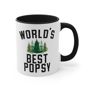 May include: White ceramic mug with a black rim and handle. The mug has a black graphic that says "World's Best Popsy" with a green watercolor illustration of pine trees.