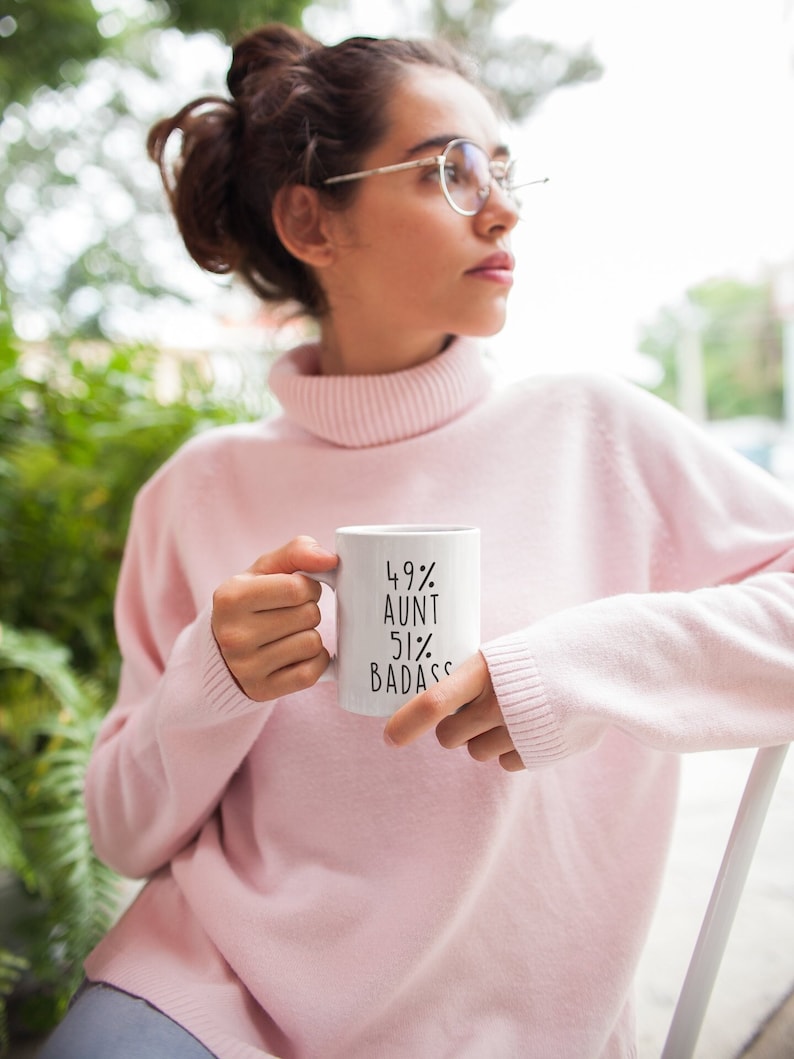 Puede incluir: Taza de cer&aacute;mica blanca con texto negro que dice "49% T&iacute;a 51% Badass". Una persona est&aacute; sosteniendo la taza.
