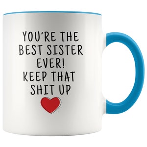 May include: White ceramic coffee mug with a blue rim and handle. The mug has black text that reads "You're the best sister ever! Keep that shit up" with a red heart below the text.