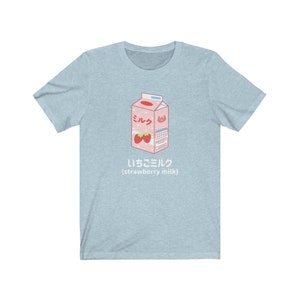 May include: A light blue t-shirt with a cartoon illustration of a carton of strawberry milk. The carton is pink and white with the Japanese word for milk, "ミルク", and the text "いちごミルク (strawberry milk)".