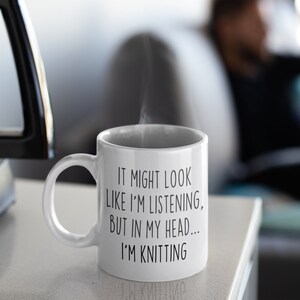 May include: White ceramic coffee mug with black text that reads "It might look like I'm listening, but in my head... I'm knitting".