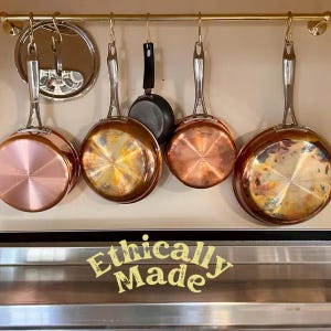 Puede incluir: Cinco ollas de color cobre cuelgan de un estante de color dorado. Las ollas son de diferentes tamaños y tienen un acabado brillante. El texto "Ethically Made" está impreso en una superficie de acero inoxidable debajo de las ollas.