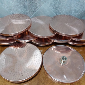 May include: A stack of 10 copper shower heads with a hammered finish. The shower heads are round and have a hole in the centre. The shower heads are stacked on top of each other, with the top shower head being the largest and the bottom shower head being the smallest.