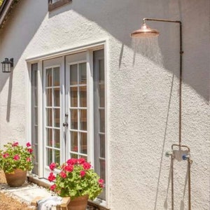 May include: A copper outdoor shower head attached to a pipe on a white stucco wall. The shower head is positioned over a small patio area with potted plants and a wooden stool.