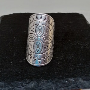 May include: Silver statement ring with an engraved geometric design. The ring has an elongated oval shape with symmetrical patterns, including floral and linear elements. The ring is displayed on a dark, textured surface.